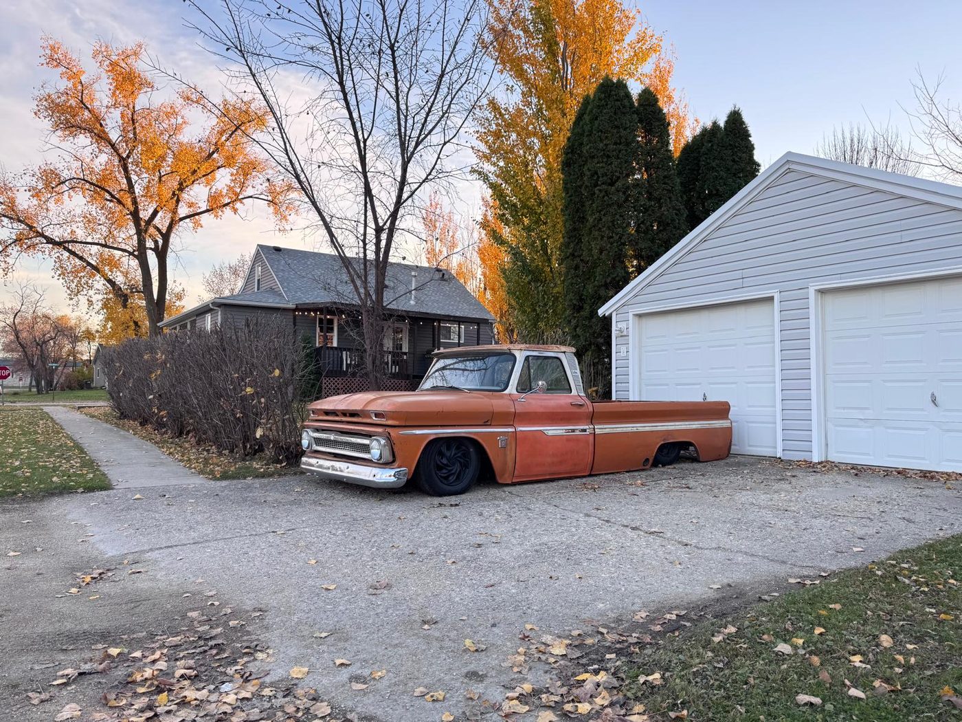 Lowered orange pickup with white roof parked in a driveway beside a garage, autumn trees behind